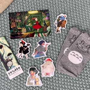 May include: A collection of Studio Ghibli themed items, including a postcard, a bookmark, stickers, and socks. The postcard features a scene from the movie "My Neighbor Totoro", the bookmark features Totoro, and the stickers feature characters from various Ghibli films. The socks are gray with a white Totoro design.