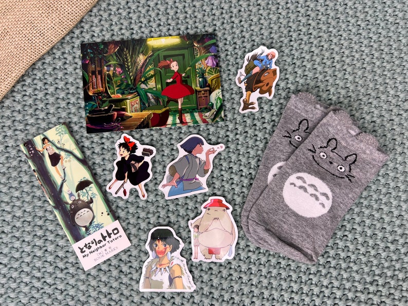 May include: A collection of Studio Ghibli themed items, including a postcard, a bookmark, stickers, and socks. The postcard features a scene from the movie "My Neighbor Totoro", the bookmark features Totoro, and the stickers feature characters from various Ghibli films. The socks are gray with a white Totoro design.
