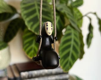May include: A black No Face figurine from the movie Spirited Away is on a swing. The figurine has a white face with purple eyes and a smile. The swing is made of brown rope.