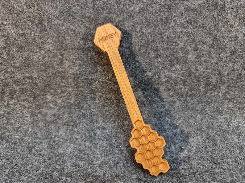 Puede incluir: Un cuchar&oacute;n de miel de madera con un dise&ntilde;o de panal y la palabra "Honey" grabada en la parte superior.