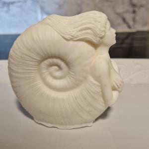 May include: White ceramic sculpture of a woman with flowing hair, sitting inside a seashell.