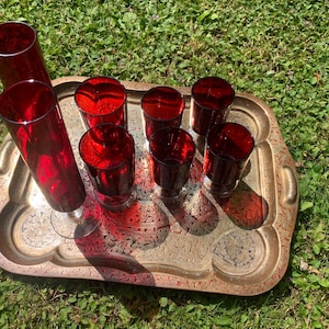 May include: A set of deep red glass drinkware consisting of one tall stemmed flute and five matching tumblers, all arranged on a rectangular brass-toned metal tray featuring an intricate engraved floral pattern across its surface.