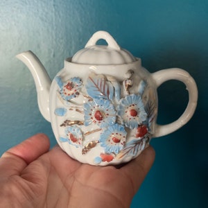 May include: A small, white ceramic teapot with a lid and handle. The teapot is decorated with blue and red floral designs and gold accents. The teapot is held in a hand.