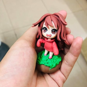 May include: A small, handmade figurine of a girl with long brown hair, wearing a red sweater and standing on a green base.