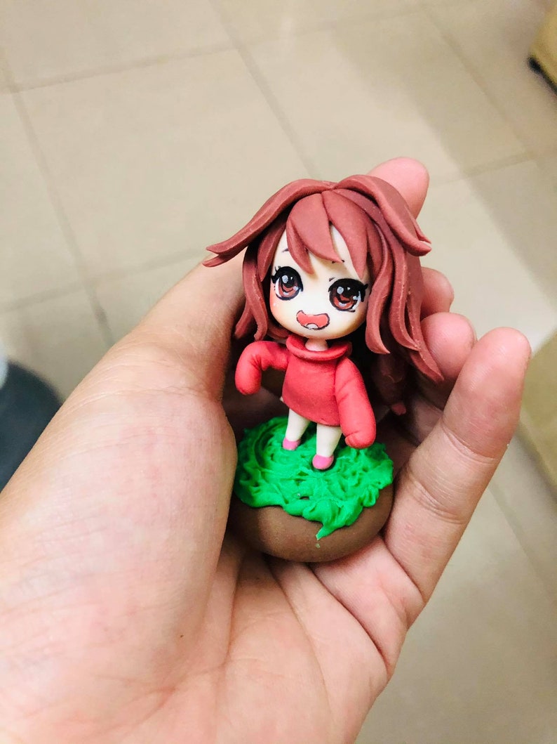 May include: A small, handmade figurine of a girl with long brown hair, wearing a red sweater and standing on a green base.