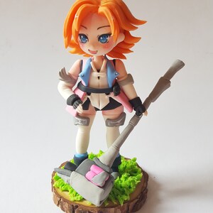 May include: A handmade figurine of a female character with orange hair, blue eyes, and a pink and blue outfit. She is holding a large grey hammer with pink accents and is standing on a wooden base with green grass.