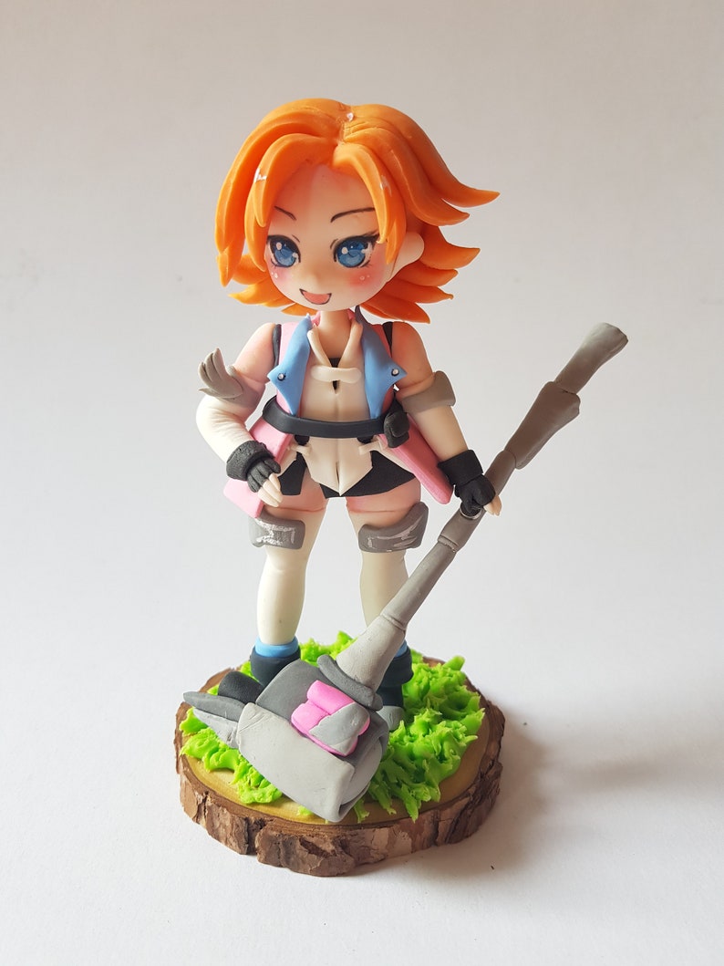May include: A handmade figurine of a female character with orange hair, blue eyes, and a pink and blue outfit. She is holding a large grey hammer with pink accents and is standing on a wooden base with green grass.