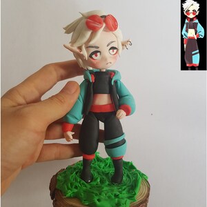 May include: A handmade polymer clay figurine of a character with white hair, red eyes, and red and black clothing. The figurine is wearing a turquoise jacket and red and black trousers. The figurine is standing on a green and brown base.
