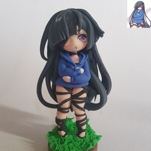 May include: A handmade figurine of a girl with long black hair, wearing a blue hoodie and black leg wraps. She has purple eyes and a light pink blush on her cheeks. The figurine is standing on a bed of green grass.