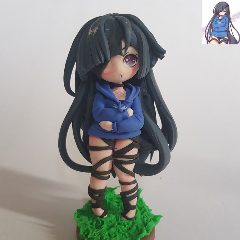 May include: A handmade figurine of a girl with long black hair, wearing a blue hoodie and black leg wraps. She has purple eyes and a light pink blush on her cheeks. The figurine is standing on a bed of green grass.