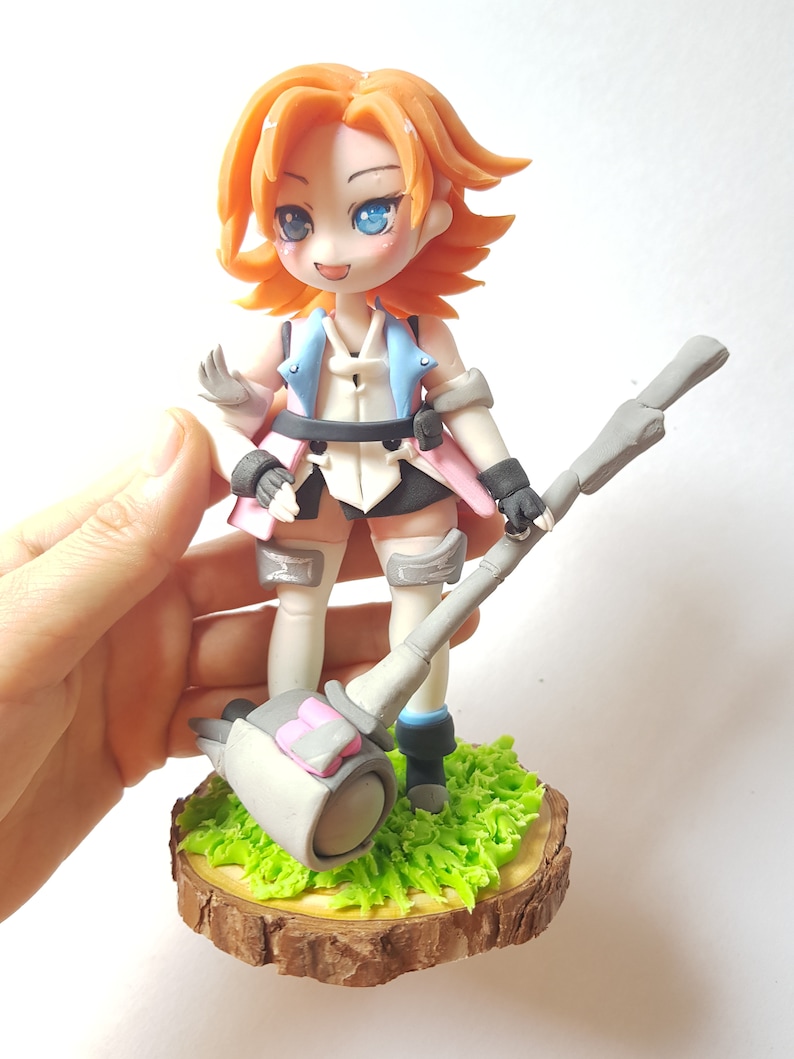 May include: A handmade figurine of a female character with orange hair, blue eyes, and a pink and blue outfit. She is holding a large grey hammer with a pink detail. The figurine is standing on a wooden base with green grass.