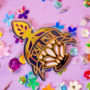 Può includere: Un adesivo argentato con una corona dorata e la parola "POUR EUX" in lettere nere. L'adesivo è circondato da paillettes e perline colorate su uno sfondo viola.