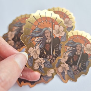 May include: Gold foil sticker featuring a woman with long blue hair holding a sword. She is wearing armor and a red heart on her chest. The background is a golden sun with white flowers.