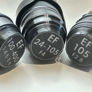 Può includere: Tre tappi per obiettivi di fotocamera neri con testo bianco. I tappi riportano "EF 100-400 1:4.5-5.6", "EF 24-105 F4" e "EF 105 F2.8".