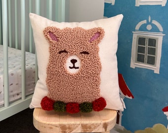 May include: A plush brown llama pillow with a white face and closed eyes. The pillow is decorated with red and green pom-poms around the bottom.