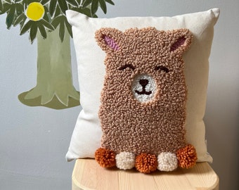 May include: A brown and beige plush pillow with a llama face and orange and white pom-poms. The pillow is sitting on a wooden stool.