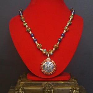 May include: A beaded necklace with a pendant displayed on a red velvet bust. The necklace features red, blue, and turquoise beads, with a silver-toned pendant. The pendant has a blue center with a silver design and is surrounded by red beads.