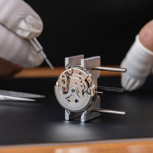 May include: A watchmaker uses tweezers to work on a silver watch movement held in a metal clamp. The watch movement is intricate and detailed, with many small gears and springs.