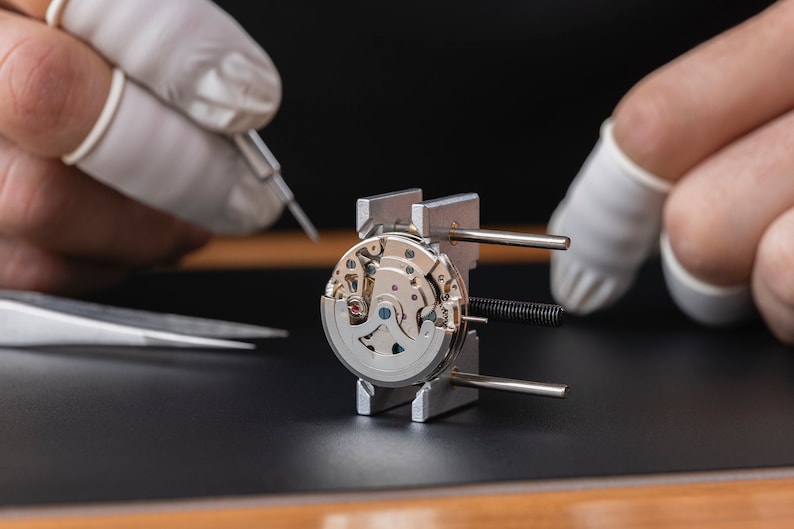 May include: A watchmaker uses tweezers to work on a silver watch movement held in a metal clamp. The watch movement is intricate and detailed, with many small gears and springs.