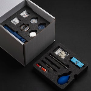 May include: A white box containing a blue watch strap and watch components. A second box with watch repair tools and a blue bulb is open on a black surface.