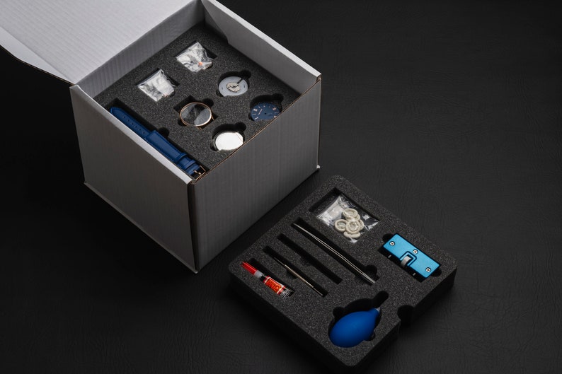May include: A white box containing a blue watch strap and watch components. A second box with watch repair tools and a blue bulb is open on a black surface.
