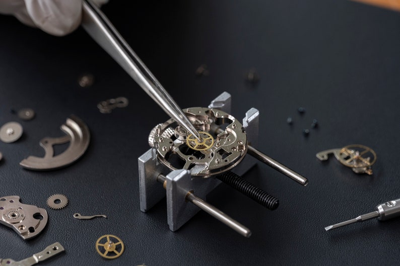 May include: A watchmaker uses tweezers to place a small, gold-colored gear into a watch movement held in a metal clamp. The watch movement is surrounded by other watch parts on a black surface.