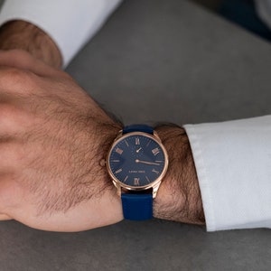 May include: A rose gold wristwatch with a blue leather strap and a blue face with Roman numeral markings. The watch has a small second hand dial at the 6 o'clock position.
