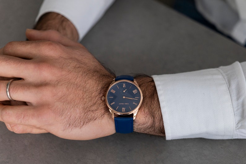 May include: A rose gold wristwatch with a blue leather strap and a blue face with Roman numeral markings. The watch has a small second hand dial at the 6 o'clock position.
