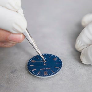 May include: A close-up of a watch face being assembled. The watch face is blue with Roman numeral hour markers and a silver hand. The watchmaker is using tweezers to place the hand on the face.
