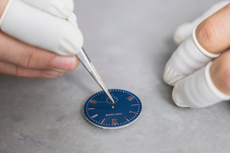 May include: A close-up of a watch face being assembled. The watch face is blue with Roman numeral hour markers and a silver hand. The watchmaker is using tweezers to place the hand on the face.