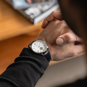 May include: A silver wristwatch with a white face and black leather strap. The watch has Roman numeral hour markers and a small second hand.