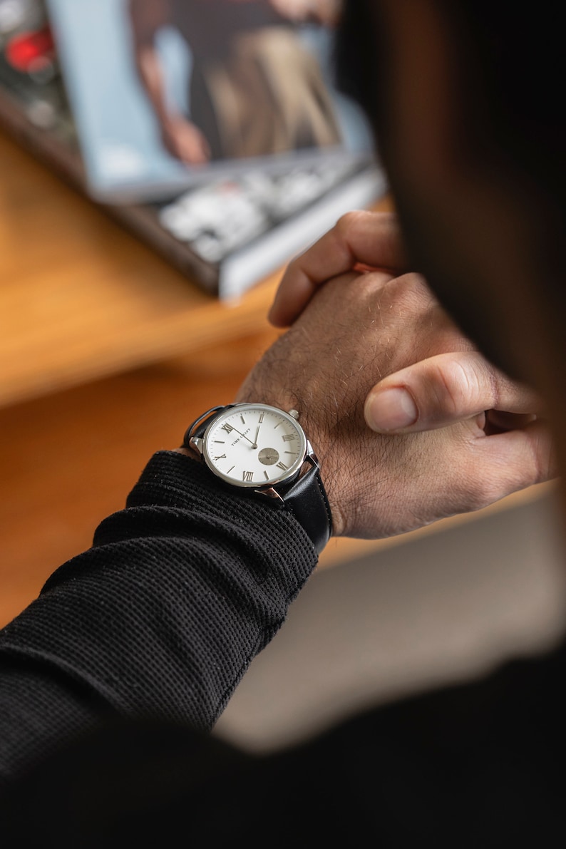 May include: A silver wristwatch with a white face and black leather strap. The watch has Roman numeral hour markers and a small second hand.