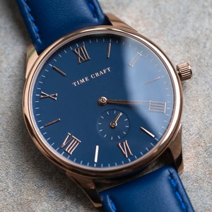 May include: A rose gold wristwatch with a blue leather strap and a blue face with Roman numeral hour markers. The watch face says "TIME CRAFT".
