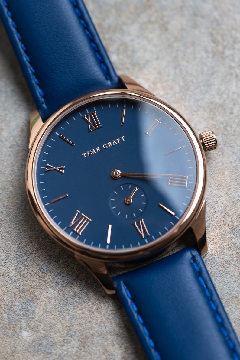 May include: A rose gold wristwatch with a blue leather strap and a blue face with Roman numeral hour markers. The watch face says "TIME CRAFT".