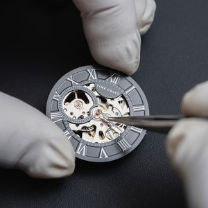 May include: A close-up of a watchmaker using tweezers to adjust the gears of a silver skeleton watch face with Roman numeral markings. The watch face reads "TIME CRAFT".