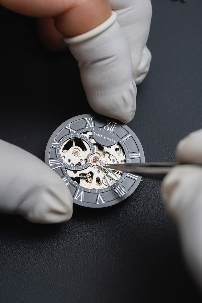 May include: A close-up of a watchmaker using tweezers to adjust the gears of a silver skeleton watch face with Roman numeral markings. The watch face reads "TIME CRAFT".