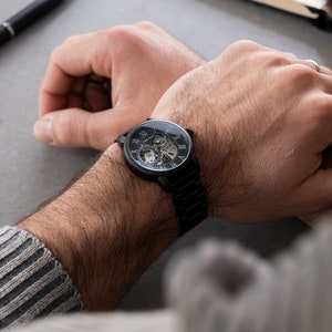 May include: A black wristwatch with a skeleton dial and Roman numeral hour markers. The watch has a black leather band.