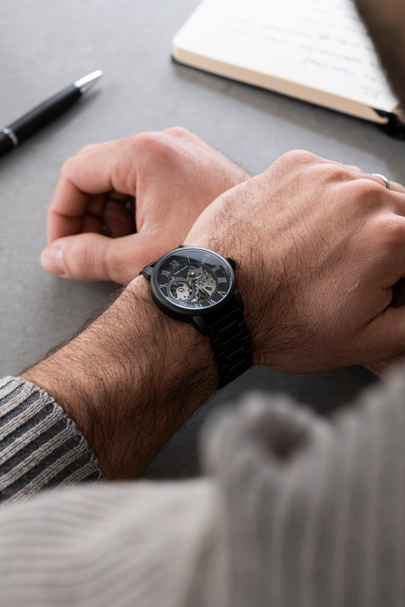 May include: A black wristwatch with a skeleton dial and Roman numeral hour markers. The watch has a black leather band.
