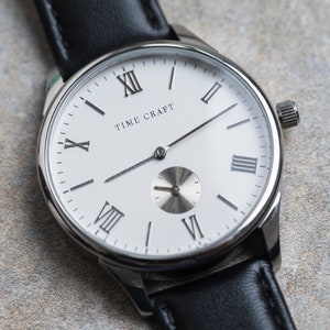 May include: A silver watch with a white face and black leather strap. The watch face has Roman numeral hour markers and the text "TIME CRAFT" printed on it.