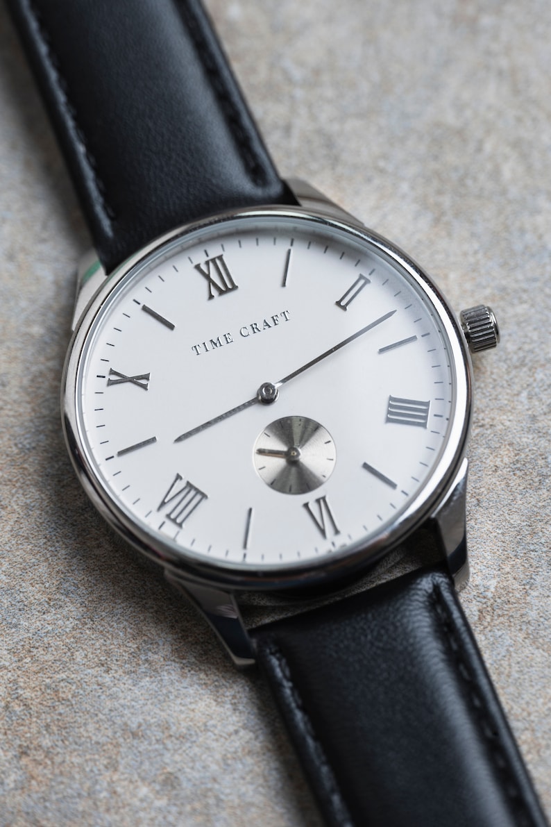 May include: A silver watch with a white face and black leather strap. The watch face has Roman numeral hour markers and the text "TIME CRAFT" printed on it.
