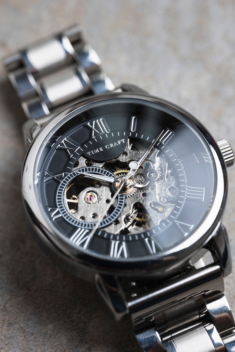 May include: A silver watch with a black face and Roman numeral hour markers. The watch has a skeletonized design, revealing the inner workings of the movement. The watch face also features the text "TIME CRAFT".