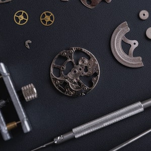 May include: A close-up of a watchmaker's workbench with various watch parts, including gears, a watch movement, and a screwdriver with a red handle.