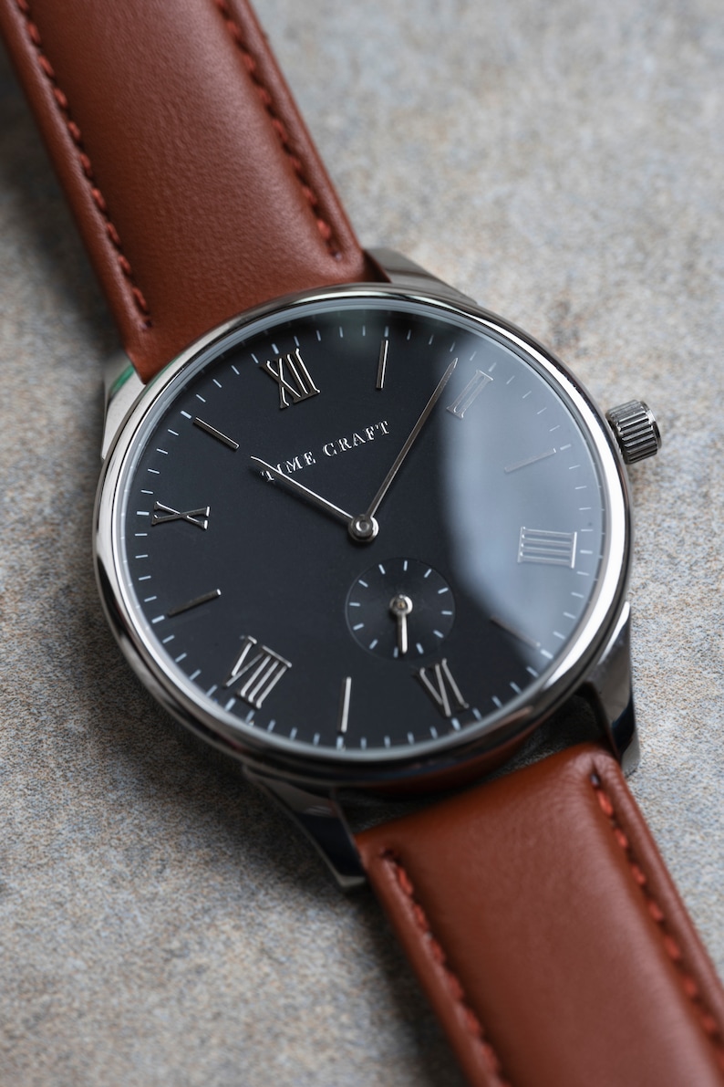 May include: A silver watch with a black face and Roman numeral hour markers. The watch has a brown leather strap and the words "Time Craft" are printed on the face.