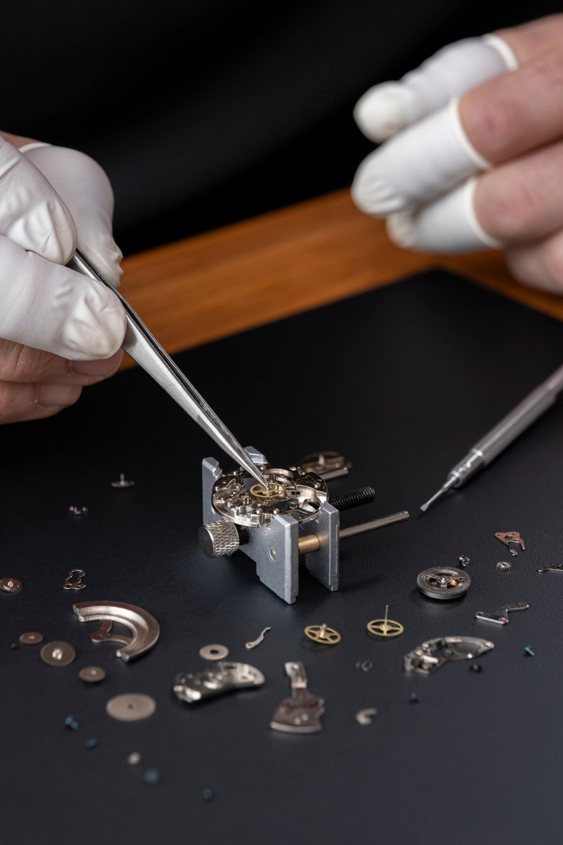 May include: A watchmaker uses tweezers to work on a watch movement, surrounded by disassembled watch parts on a black surface.
