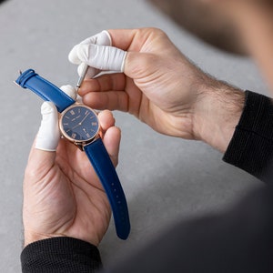 May include: A person wearing white gloves adjusts the strap of a rose gold watch with a blue leather band and a blue face.