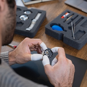 May include: A person is working on a watch face, holding it with two fingers covered in white gloves. The watch face is silver with black Roman numeral markings and a black background.