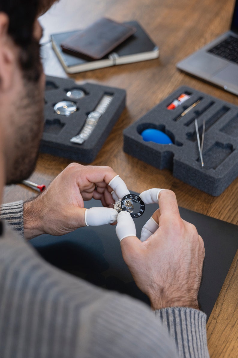 May include: A person is working on a watch face, holding it with two fingers covered in white gloves. The watch face is silver with black Roman numeral markings and a black background.