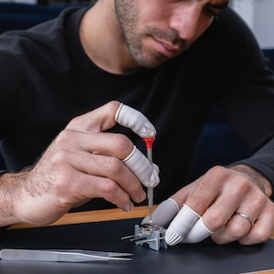 May include: A person wearing white finger cots uses a small screwdriver to work on a silver watch mechanism. The watch mechanism is on a black surface with a silver tweezers nearby.