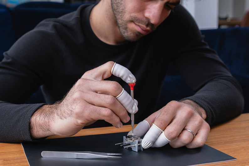 May include: A person wearing white finger cots uses a small screwdriver to work on a silver watch mechanism. The watch mechanism is on a black surface with a silver tweezers nearby.