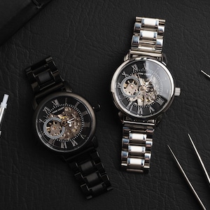 May include: Two skeleton watches with black faces and Roman numeral hour markers. The watch on the left has a black metal band and the watch on the right has a silver metal band. Both watches are on a black leather surface.